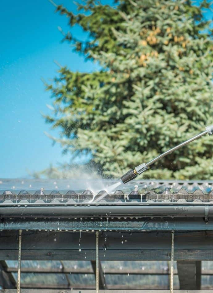 Pressure washer cleaning gutters, removing debris. Clear blue sky and green tree in background. Scarborough ME homeowners maintaining their gutters to prevent water damage.