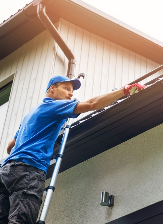 Man on ladder cleaning gutters of a house, wearing a blue shirt and cap. He is removing debris from the gutter, ensuring proper drainage. Homeowners seeking "Gutter Cleaning Near Me Falmouth ME" can rely on professionals for maintenance.